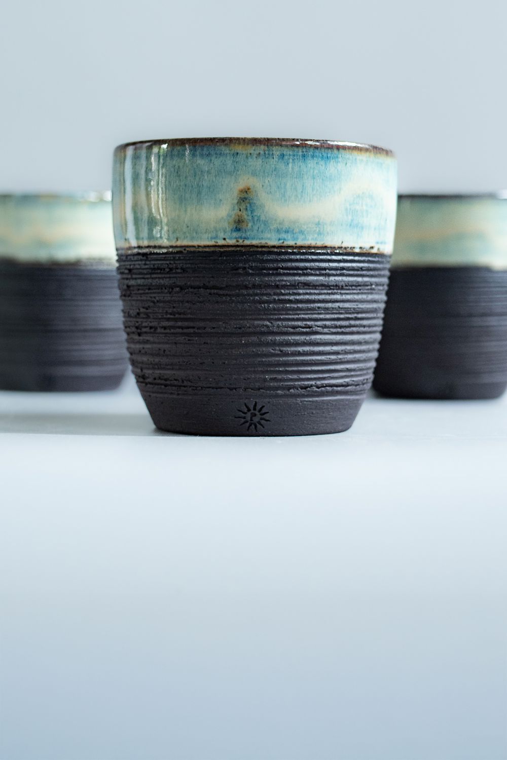 Textured blue and black ceramic coffee cup by Potterbeans with a rustic glazed finish centered against a pale background with two matching cups behind it