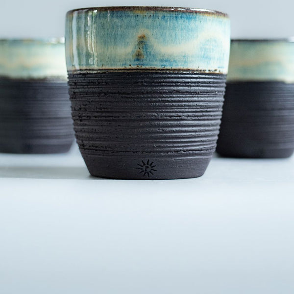 Textured blue and black ceramic coffee cup by Potterbeans with a rustic glazed finish centered against a pale background with two matching cups behind it